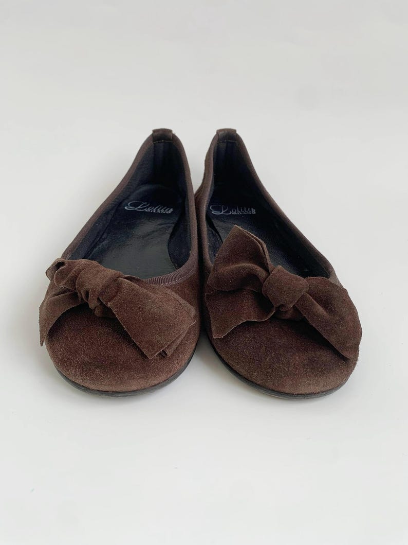 May include: A pair of brown suede ballet flats with large bow accents. The shoes have a rounded toe and a flat sole. The brand name "Lalita" is visible on the insole. The shoes are set against a white background.