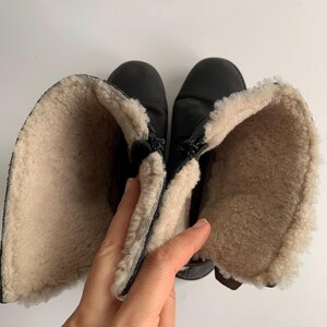 May include: A pair of black boots featuring a thick, cream-coloured fleece lining. The boots are shown from a top-down perspective with the shafts folded open to reveal the interior texture and the black exterior material of the footwear.