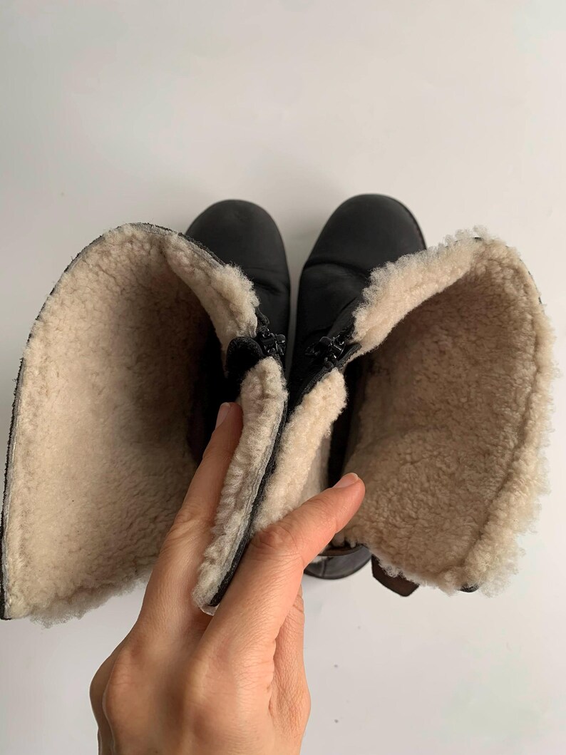 May include: A pair of black boots featuring a thick, cream-coloured fleece lining. The boots are shown from a top-down perspective with the shafts folded open to reveal the interior texture and the black exterior material of the footwear.