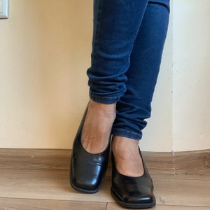 May include: A pair of black leather square-toe flats. The shoes have a low heel and a smooth, shiny finish. The flats are worn with dark blue denim jeans.
