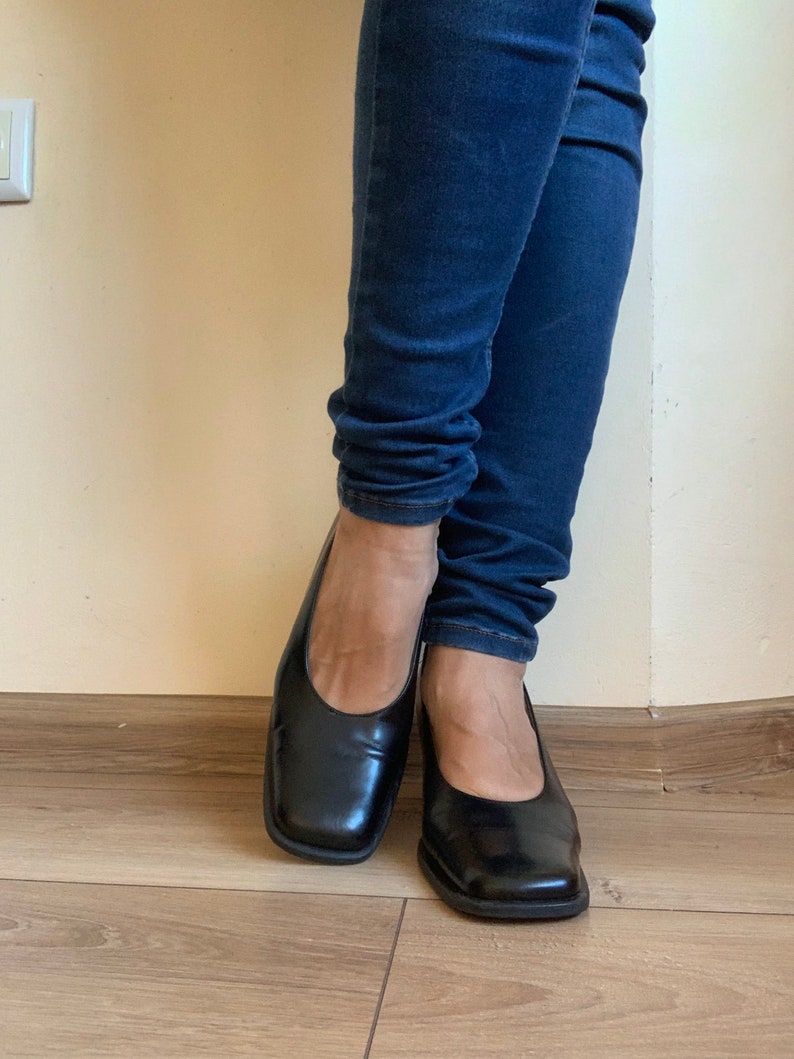 May include: A pair of black leather square-toe flats. The shoes have a low heel and a smooth, shiny finish. The flats are worn with dark blue denim jeans.