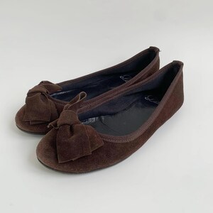 May include: A pair of brown suede ballet flats. Each flat features a bow on the toe and a rounded toe design. The shoes are a dark brown color and appear to be unworn.