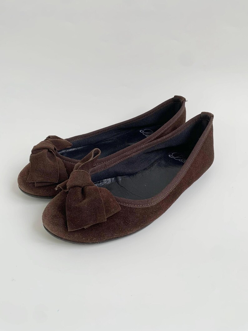 May include: A pair of brown suede ballet flats. Each flat features a bow on the toe and a rounded toe design. The shoes are a dark brown color and appear to be unworn.