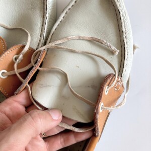 May include: A pair of white leather boat shoes with brown leather accents and laces. The shoes are worn and have some dirt and scuffs.