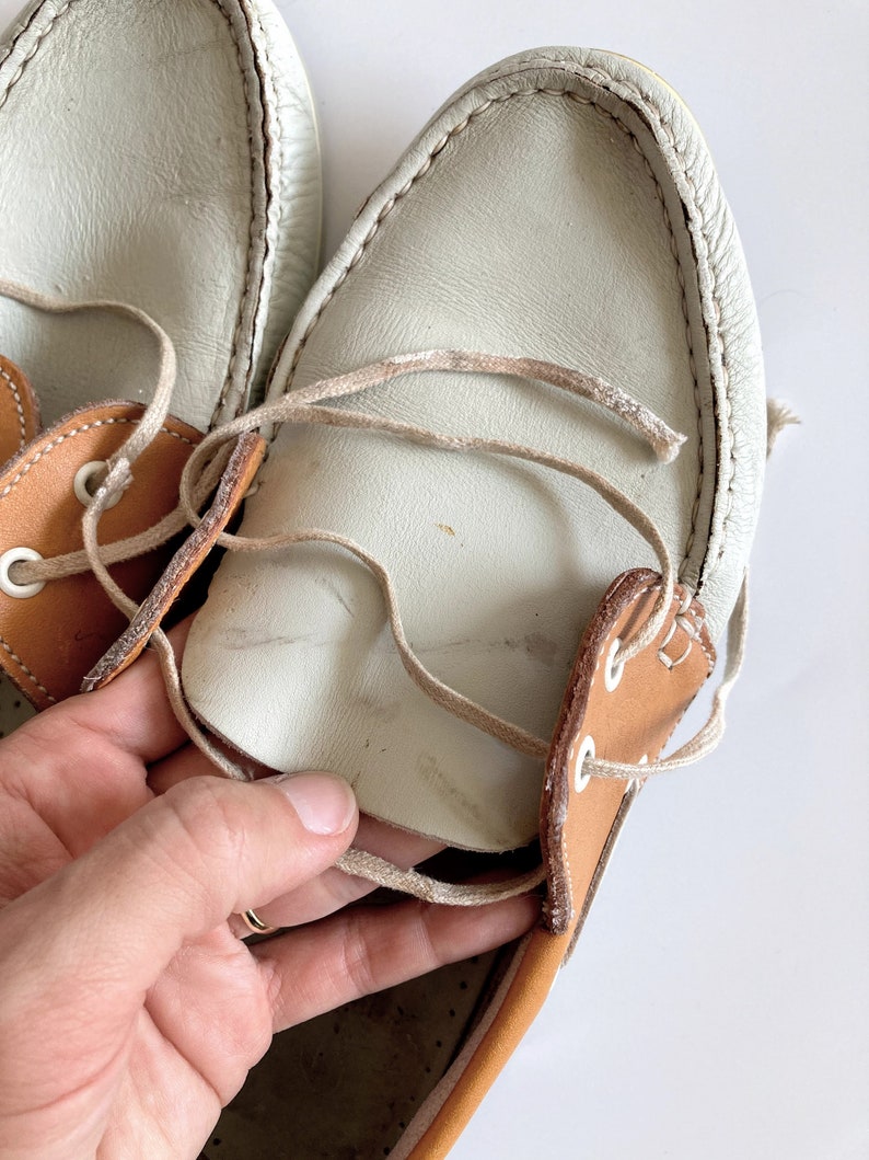 May include: A pair of white leather boat shoes with brown leather accents and laces. The shoes are worn and have some dirt and scuffs.