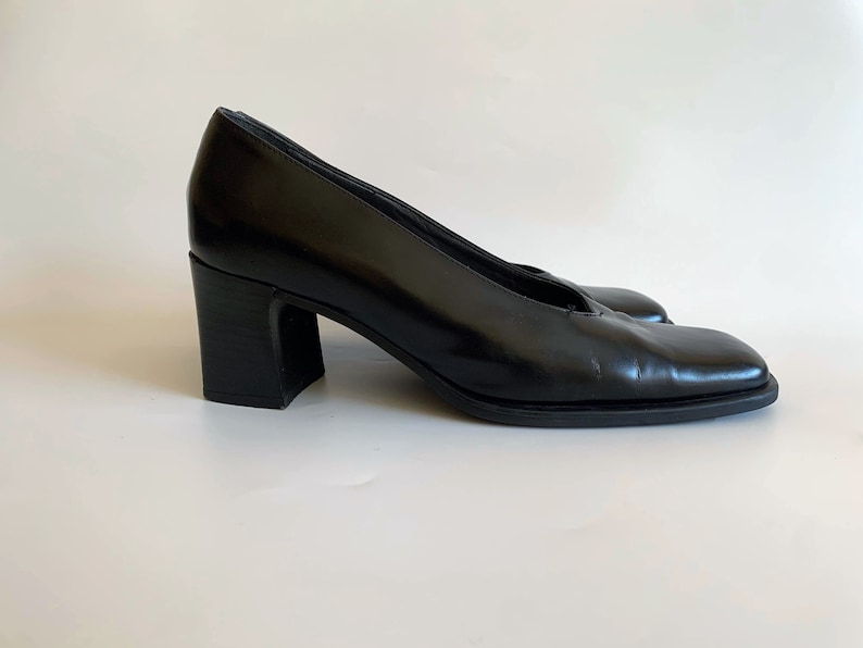 May include: A pair of black leather pumps with a square toe and a block heel. The heels are approximately 3 inches high. The shoes have a classic, minimalist design.