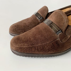 May include: A pair of brown suede loafers with black buckle accents. The loafers have a rounded toe and a low heel.
