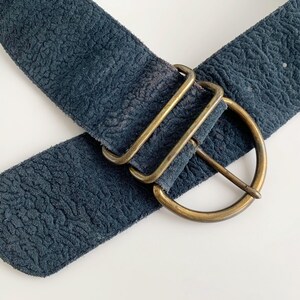 May include: A wide, navy blue belt with a textured finish. It has a large, brass-coloured buckle with a D-ring design and two rectangular keepers. The belt is angled across a white surface.