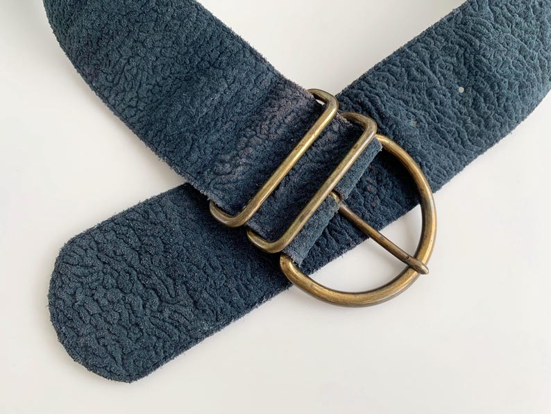 May include: A wide, navy blue belt with a textured finish. It has a large, brass-coloured buckle with a D-ring design and two rectangular keepers. The belt is angled across a white surface.