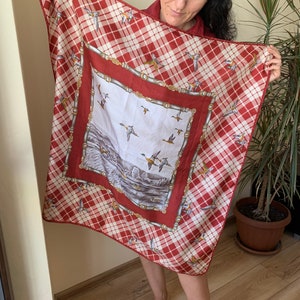 May include: A red and white plaid silk scarf with a smaller square panel featuring a scene of ducks flying over a lake. The panel is bordered with a gold chain design.