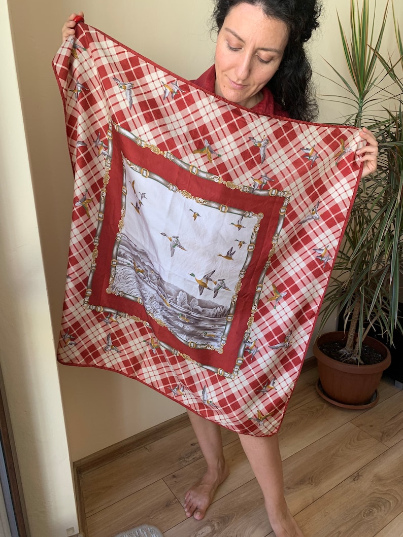 May include: A red and white plaid silk scarf with a smaller square panel featuring a scene of ducks flying over a lake. The panel is bordered with a gold chain design.