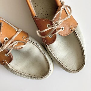 May include: A pair of white and brown leather boat shoes with laces. The shoes have a white leather upper and a brown leather trim around the top. The laces are white and are tied in a bow.
