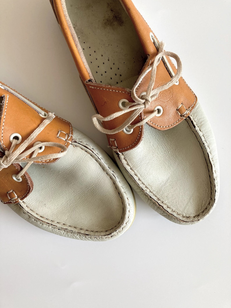 May include: A pair of white and brown leather boat shoes with laces. The shoes have a white leather upper and a brown leather trim around the top. The laces are white and are tied in a bow.