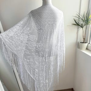 Puede incluir: Un chal de encaje blanco con flecos, exhibido en un maniquí. El chal presenta un patrón floral detallado y un tejido abierto y delicado. Los largos flecos cuelgan de los bordes, agregando movimiento y textura a la prenda. El chal es un accesorio ligero y aireado.