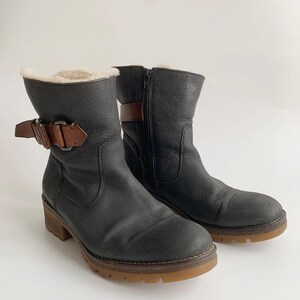 May include: A pair of black leather ankle boots featuring a textured finish, a side zip, and a brown leather strap with a metal buckle at the ankle. The boots are lined with a cream-coloured faux fur material and have a thick, brown rubber lug sole.