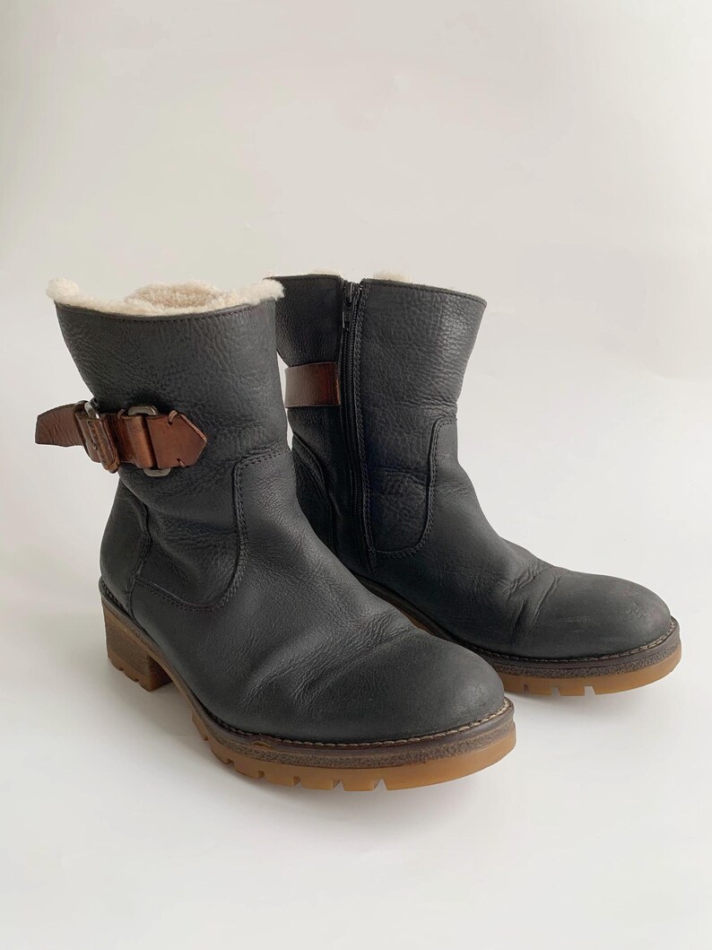 May include: A pair of black leather ankle boots featuring a textured finish, a side zip, and a brown leather strap with a metal buckle at the ankle. The boots are lined with a cream-coloured faux fur material and have a thick, brown rubber lug sole.