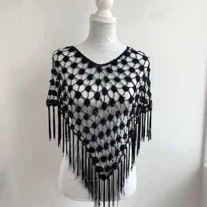 Può includere: Poncho nero all'uncinetto con scollo a V e frange. Il motivo a trama aperta crea un aspetto delicato e arioso. Il poncho è esposto su un manichino bianco, che ne mostra la forma e la consistenza.