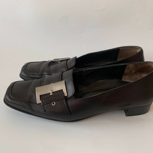 K&ouml;nnte beinhalten: Ein Paar schwarze Leder-Slipper mit einer silbernen Schnalle. Die Slipper haben eine spitze Zehenpartie und einen niedrigen Absatz.