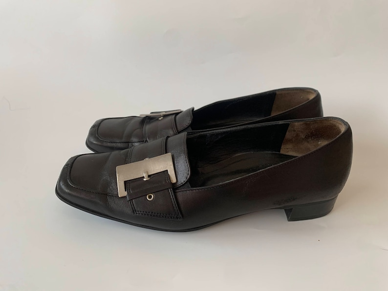 K&ouml;nnte beinhalten: Ein Paar schwarze Leder-Slipper mit einer silbernen Schnalle. Die Slipper haben eine spitze Zehenpartie und einen niedrigen Absatz.