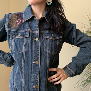 Könnte beinhalten: Eine dunkelblaue Jeansjacke mit gestickten Blumen- und Blattmotiven auf der linken Schulter. Die Jacke hat einen klassischen Stil mit Knopfleiste und langen Ärmeln.