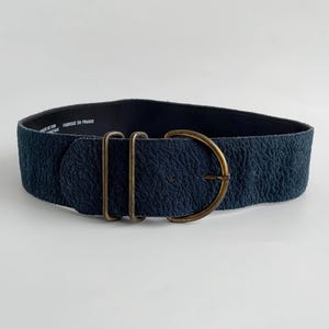 May include: A wide, navy blue belt with a textured surface and a large, antique brass buckle. The belt features two brass loops and a D-shaped buckle. The words "FABRIQUE EN FRANCE" are printed on the inside of the belt.