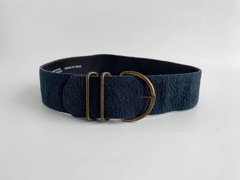 May include: A wide, navy blue belt with a textured surface and a large, antique brass buckle. The belt features two brass loops and a D-shaped buckle. The words "FABRIQUE EN FRANCE" are printed on the inside of the belt.