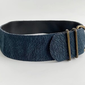 May include: A wide, navy blue belt with a textured surface and a dark interior. The belt features a brass-coloured buckle and two matching loops. The overall design is simple and elegant.