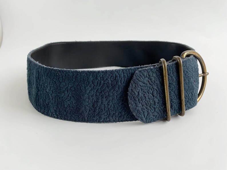 May include: A wide, navy blue belt with a textured surface and a dark interior. The belt features a brass-coloured buckle and two matching loops. The overall design is simple and elegant.