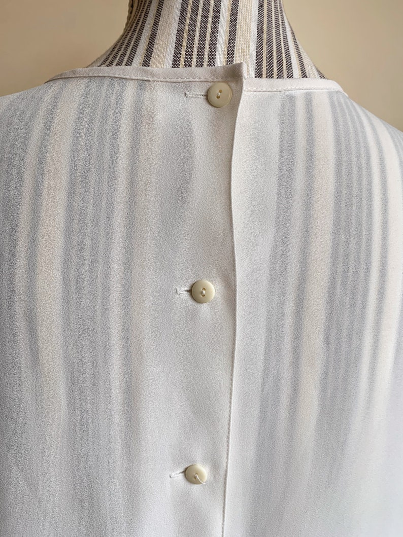 Puede incluir: Camisa blanca de botones con estampado de rayas. La camisa tiene tres botones en la espalda.