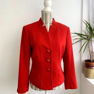 Könnte beinhalten: Ein leuchtend roter Blazer mit Stehkragen und drei passenden Knöpfen. Die maßgeschneiderte Jacke wird auf einer Schaufensterpuppe präsentiert, die ihr strukturiertes Design und die langen Ärmel hervorhebt. Ein klassisches Kleidungsstück.