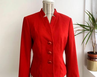 Puede incluir: Una vibrante blazer roja con cuello mandarín y tres botones a juego. La chaqueta entallada se exhibe en un maniquí, mostrando su diseño estructurado y mangas largas. Una prenda de vestir clásica.