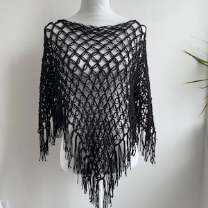 May include: Black open-weave poncho with fringe. The poncho is displayed on a white mannequin. The open weave design creates a net-like pattern. The fringe hangs from the bottom edge of the poncho. The garment is made of a soft, draping material.