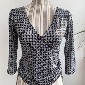 May include: Black and white patterned top with a V-neck and three-quarter sleeves. The top features a repeating geometric design and is displayed on a mannequin. The fabric appears to be a lightweight material.