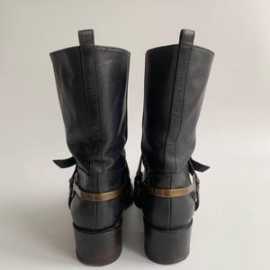 May include: A pair of black leather mid-calf boots featuring a stacked heel, a pull tab at the top, and a metallic strap across the heel area engraved with the text BOSS ORANGE. The boots have a smooth finish and a structured, upright silhouette.