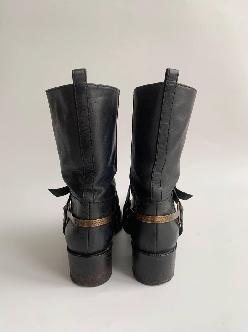 May include: A pair of black leather mid-calf boots featuring a stacked heel, a pull tab at the top, and a metallic strap across the heel area engraved with the text BOSS ORANGE. The boots have a smooth finish and a structured, upright silhouette.