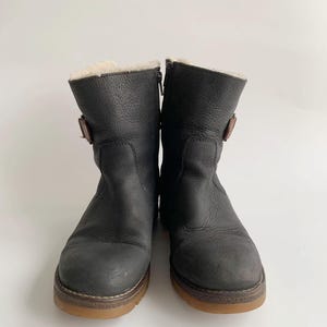 May include: A pair of mid-calf boots in dark charcoal leather with a textured finish. Each boot features a side zip, a small brown buckle strap on the outer side, and a thick gum-coloured rubber sole. The interior is lined with cream-coloured fleece.