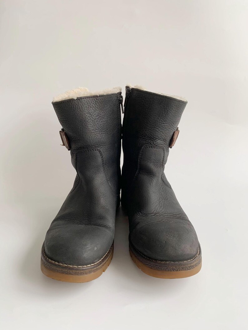May include: A pair of mid-calf boots in dark charcoal leather with a textured finish. Each boot features a side zip, a small brown buckle strap on the outer side, and a thick gum-coloured rubber sole. The interior is lined with cream-coloured fleece.