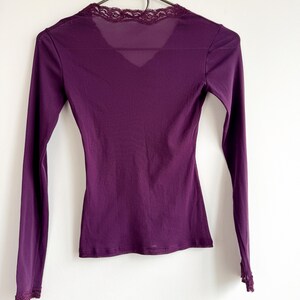 May include: A long-sleeved, deep purple top with a V-neck and delicate lace trim along the neckline and cuffs. The top is hanging on a black hanger against a white background. The fabric appears to be a soft, lightweight material.