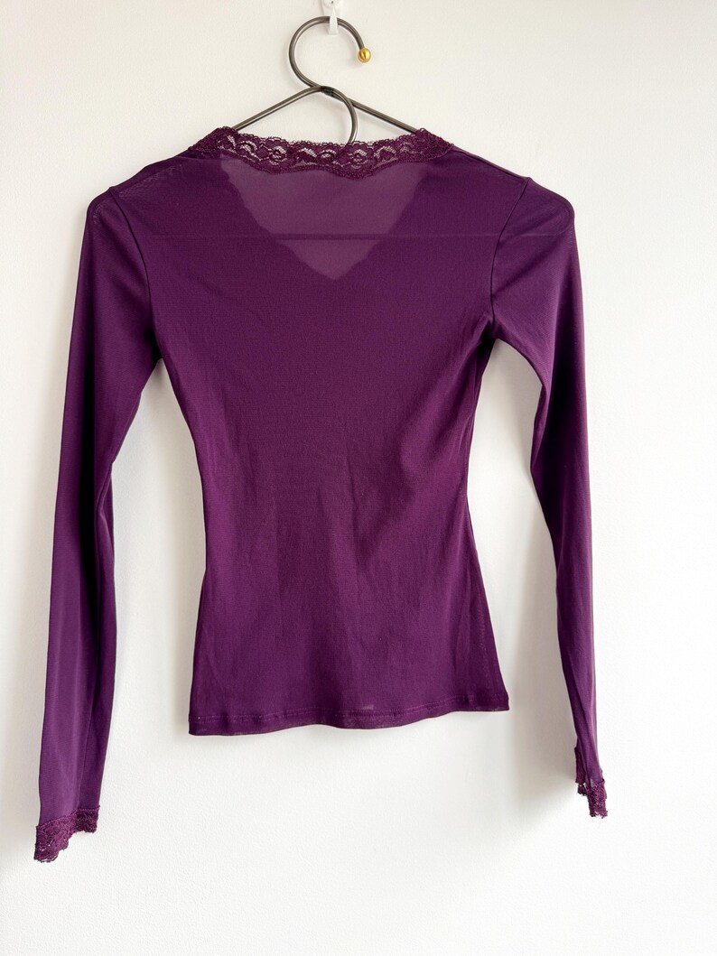 May include: A long-sleeved, deep purple top with a V-neck and delicate lace trim along the neckline and cuffs. The top is hanging on a black hanger against a white background. The fabric appears to be a soft, lightweight material.