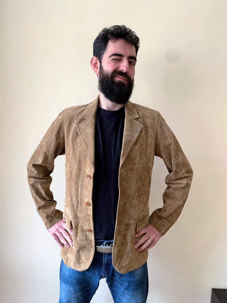 May include: A brown suede jacket with a button closure and two pockets. The jacket is worn over a black t-shirt and blue jeans.