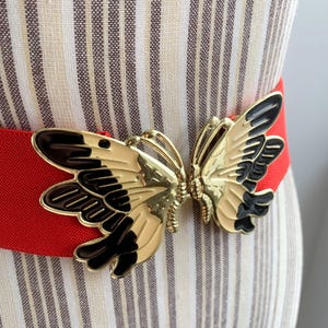 Könnte beinhalten: Ein roter elastischer Gürtel mit einer goldfarbenen Schmetterlingsschnalle. Der Schmetterling hat schwarze und cremefarbene Akzente. Der Gürtel wird auf einer Schaufensterpuppe mit gestreiftem Stoffhintergrund präsentiert.