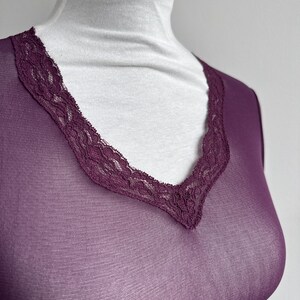 May include: A sheer, long-sleeved, plum-colored top with a V-neckline. The neckline is trimmed with a delicate, dark purple lace. The top is displayed on a white mannequin, showcasing the garment's shape and design.