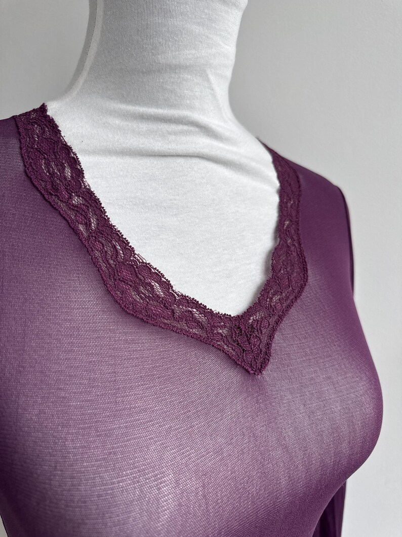 May include: A sheer, long-sleeved, plum-colored top with a V-neckline. The neckline is trimmed with a delicate, dark purple lace. The top is displayed on a white mannequin, showcasing the garment's shape and design.