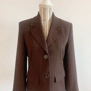 May include: A brown blazer with thin white stripes. The blazer has a notched collar, two button closure, and a single pocket on the left side.