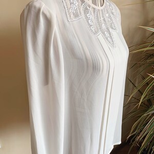 Puede incluir: Una blusa blanca de gasa con un delicado cuello de encaje. La blusa tiene mangas largas y un escote ligeramente fruncido.