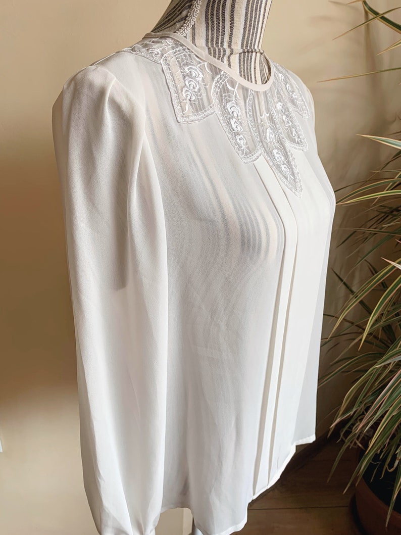 Puede incluir: Una blusa blanca de gasa con un delicado cuello de encaje. La blusa tiene mangas largas y un escote ligeramente fruncido.