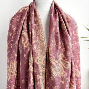 May include: A draped, burgundy scarf with a paisley pattern in beige and light brown. The scarf is displayed on a white mannequin and features a detailed floral design. The fabric appears soft and flowing, suitable for a shawl or wrap.