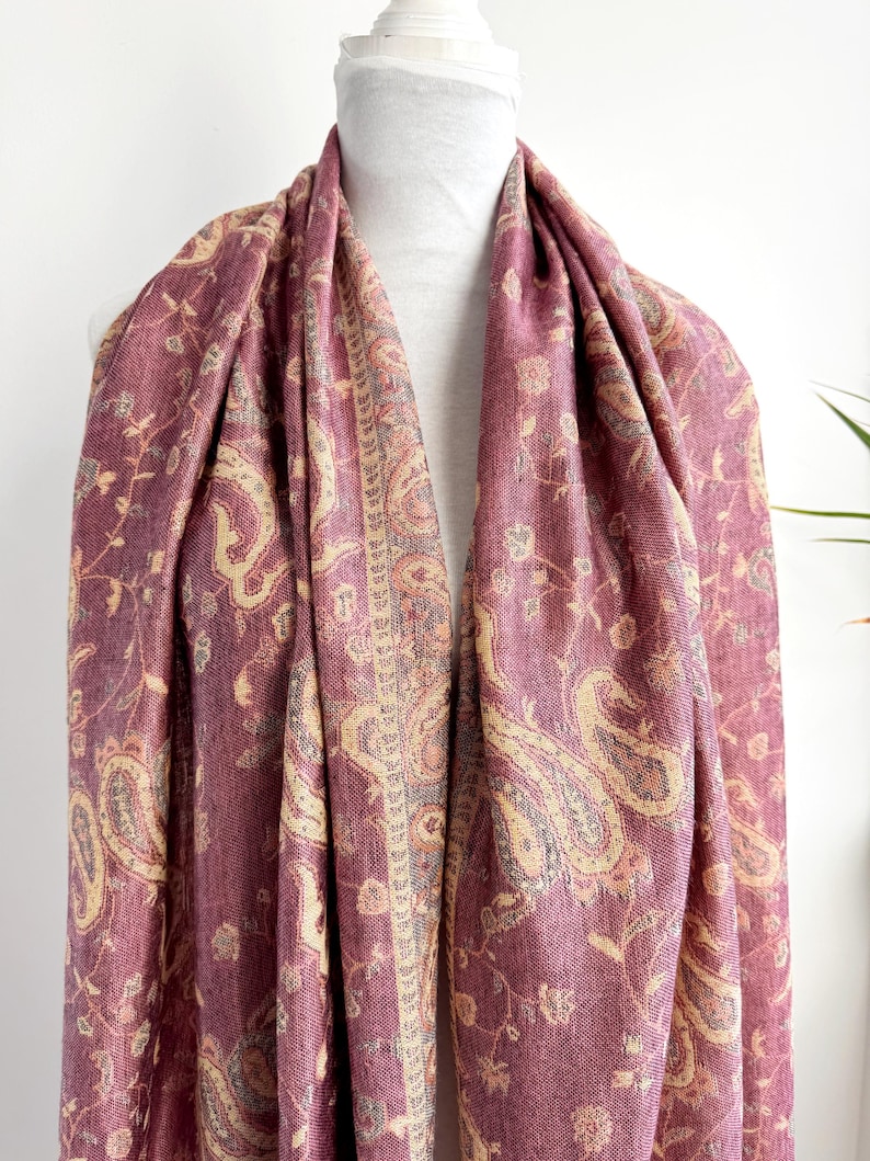 May include: A draped, burgundy scarf with a paisley pattern in beige and light brown. The scarf is displayed on a white mannequin and features a detailed floral design. The fabric appears soft and flowing, suitable for a shawl or wrap.