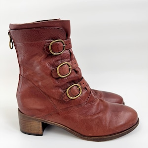 May include: Brown leather ankle boots with three brass buckles and a small heel. The boots have a zip at the back and a rounded toe. The leather has a distressed appearance.
