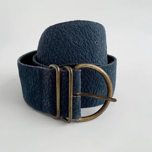 May include: A wide, navy blue belt with a textured surface. The belt features a large, antique brass buckle with a rectangular frame and a rounded buckle. The belt is approximately 7.6 cm wide.